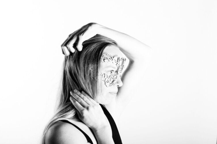 A woman with long hair poses while wearing a decorative lace mask on her face.