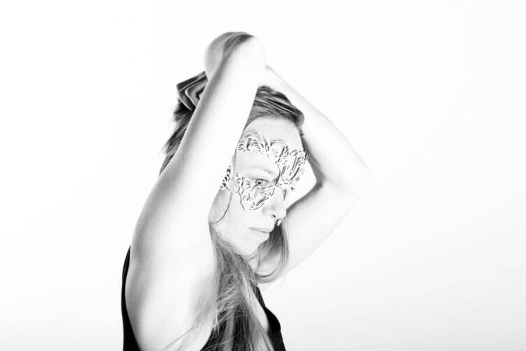 A woman with long hair wearing a decorative mask, holding her hair up in a black and white portrait.