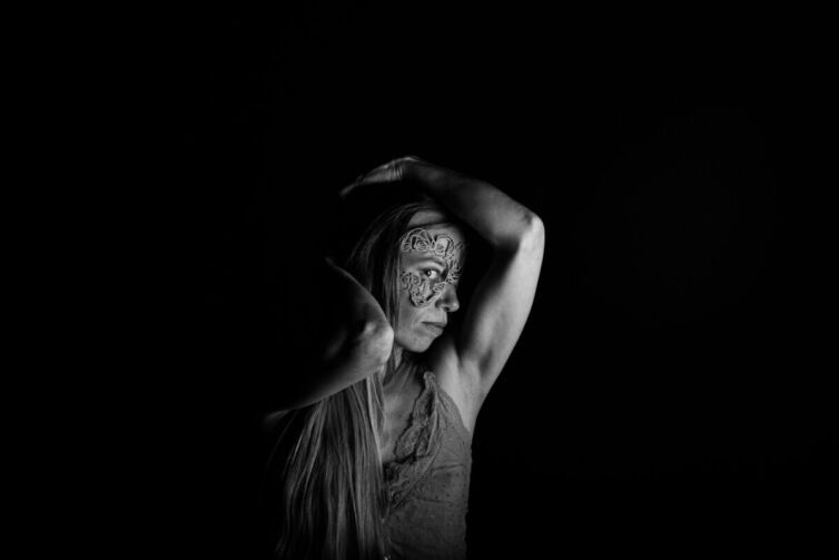 Black and white portrait of a woman with intricate face art and long hair, posing with arms raised.