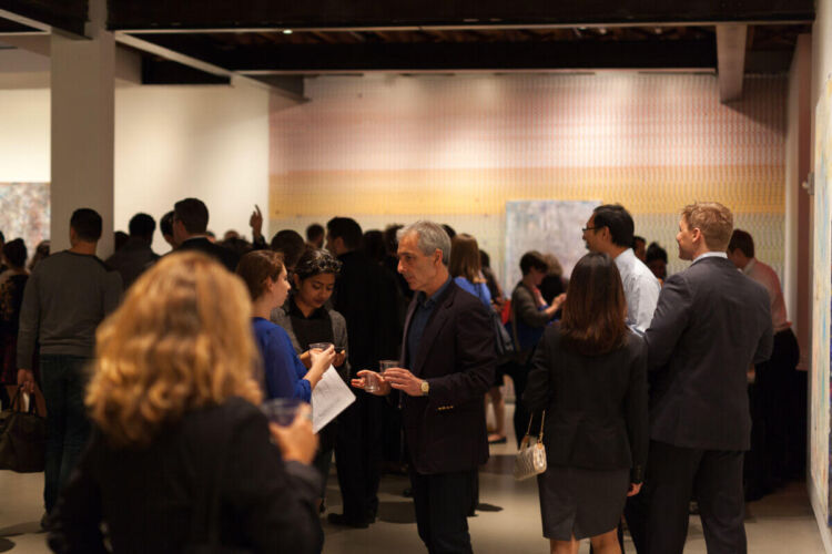 A crowded art gallery event with attendees mingling and holding drinks in a well-lit space.