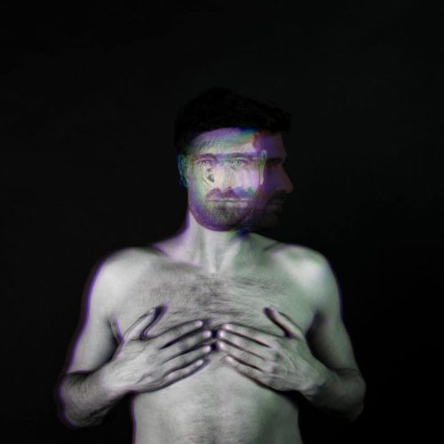 A man with a distorted face effect, standing shirtless against a dark background.
