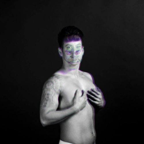 Black and white portrait of a man with tattoos, showing a playful expression and hands on his chest.