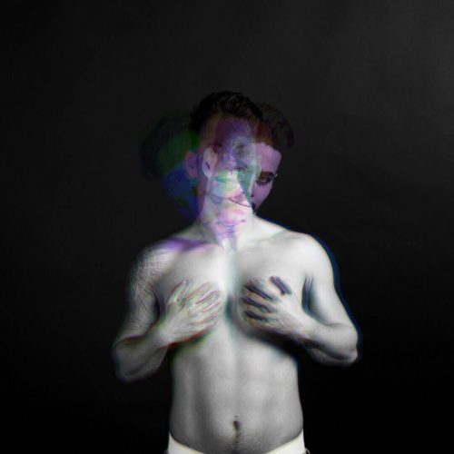 A black and white portrait of a male figure with a blurred effect, showing a torso and hands on the chest.