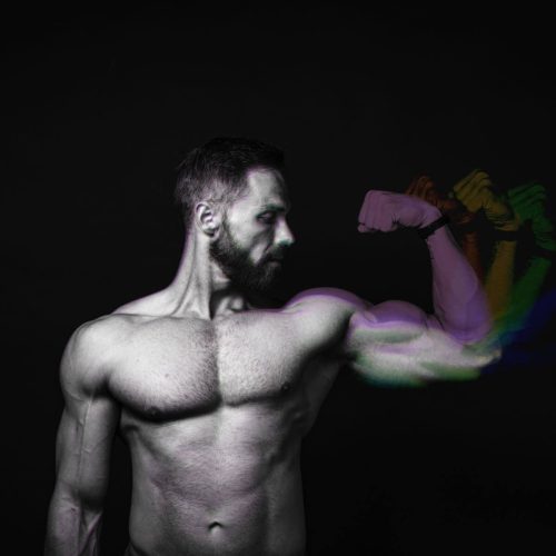 A muscular man with a beard flexing his bicep against a dark background, showing motion blur of his arm.